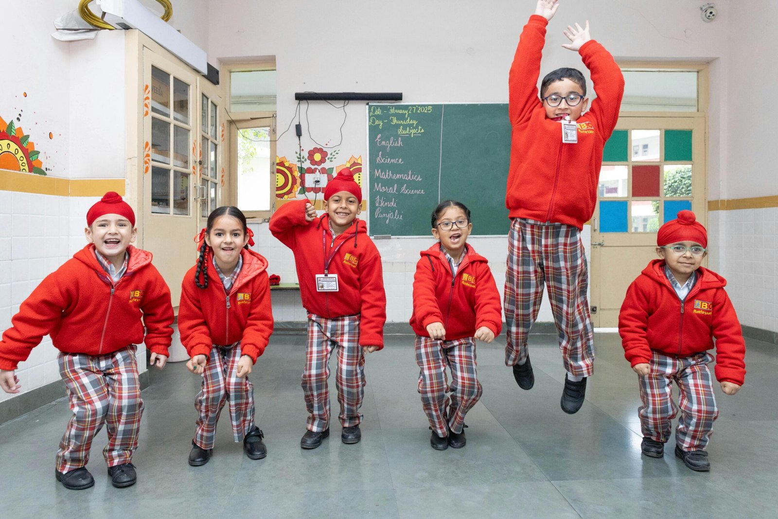 A group of young school children in red uniforms happily jumping and playing inside a colorful classroom during a school activity.